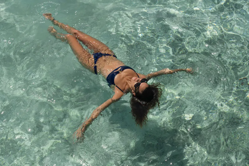 Woman in dark bikini floating relaxed on her back in clear turquoise Maldives waters, sunglasses on, arms outstretched.