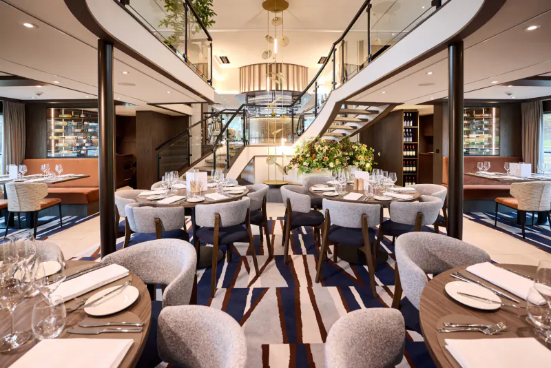 Elegant multi-level cruise ship dining room with set tables, wine racks, plants, and glass railings.