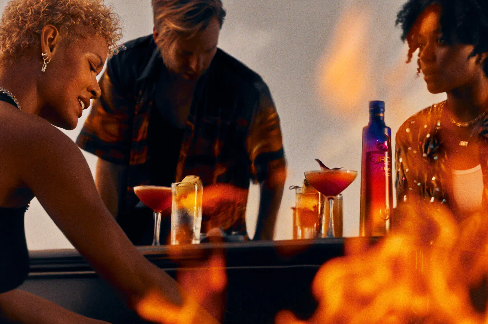 Three diverse people leaning over bar counter with CÎROC Passion vodka bottle and passion fruit cocktails amid flames