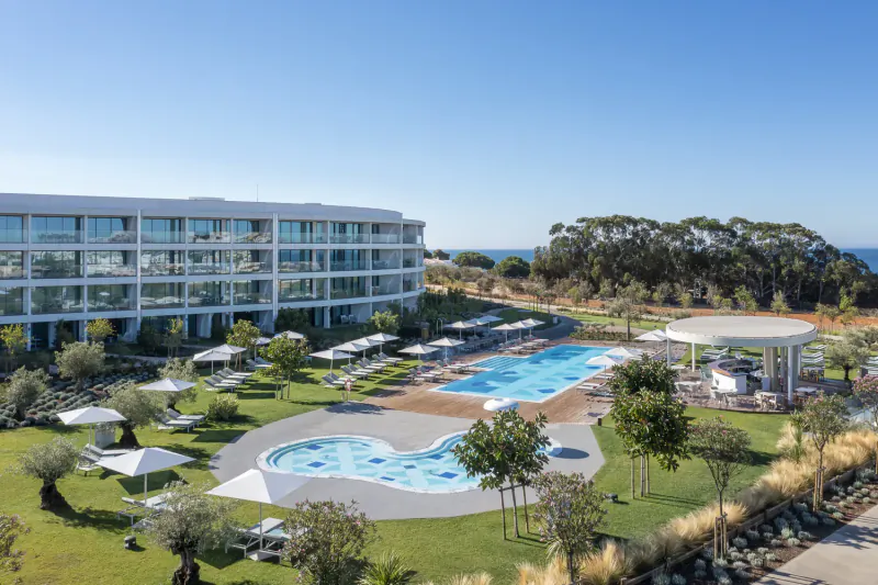 Aerial view of W Algarve Hotel with heart-shaped pool, infinity pool, white buildings, gardens, and ocean backdrop on sunny day.