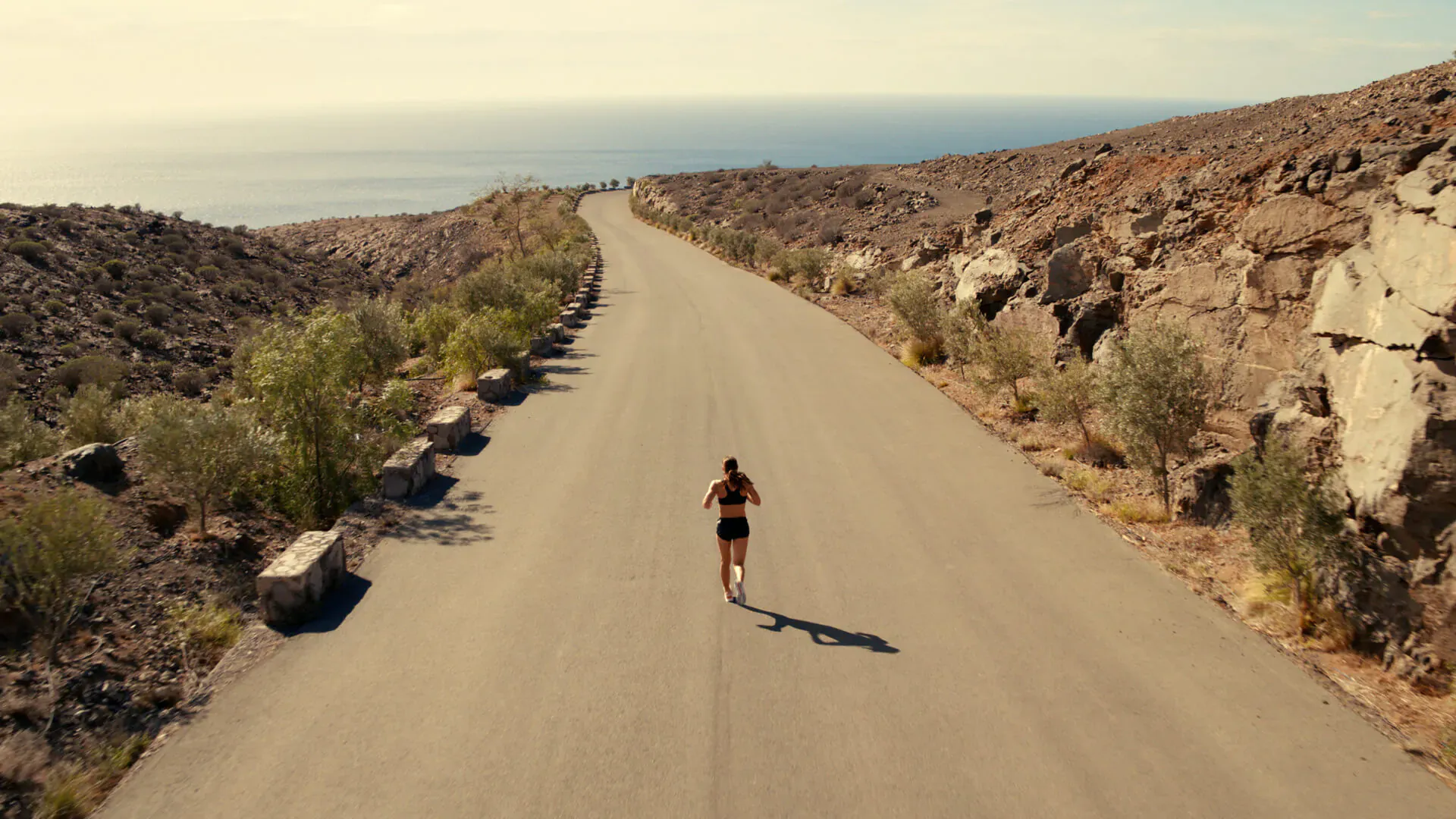 Female triathlete Nicola running on coastal mountain road, surrounded by rocks and shrubs, golden light.