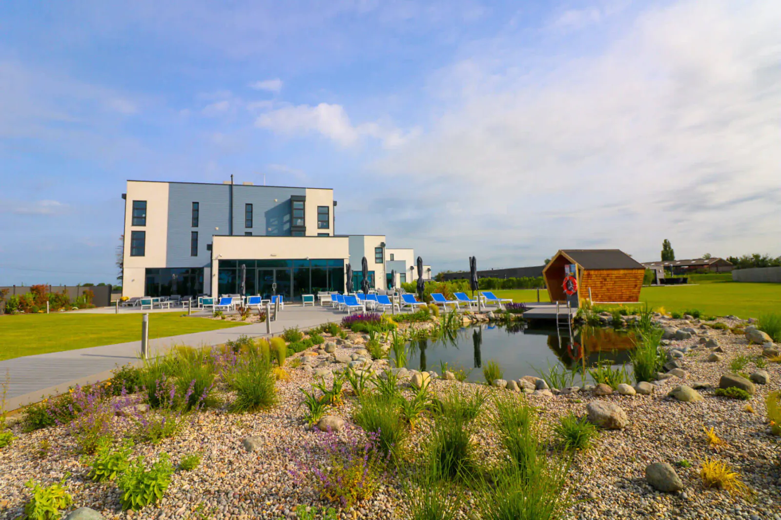 Modern white Glass House skincare retreat with blue loungers by pool, wooden cabin, koi pond, and gardens under cloudy sky