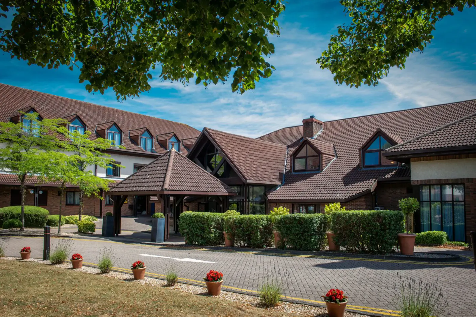 Bristol Aztec Hotel and Spa exterior with red-tiled roofs, trees, hedges, flower pots, and paved entrance under blue sky.