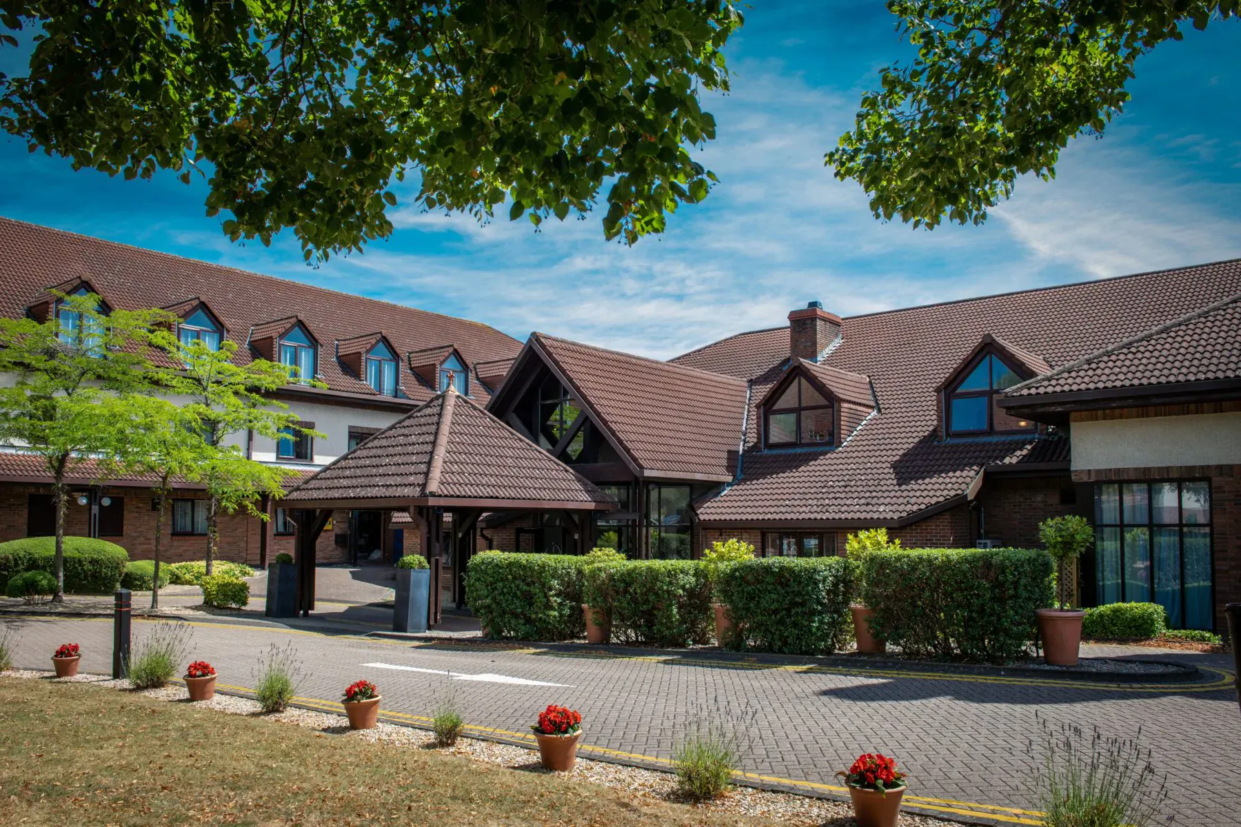 Bristol Aztec Hotel and Spa exterior with red-tiled roofs, trees, hedges, flower pots, and paved entrance under blue sky.