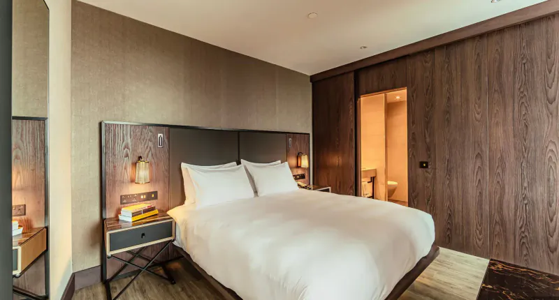 Luxury apartment bedroom at The Fellows House Cambridge with white bed, wood panels, lamps, and en-suite bathroom.