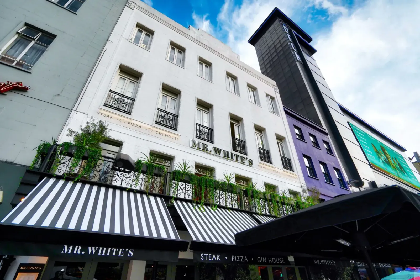 Mr White's steak pizza restaurant facade in London, white building with black striped awning, plants, and colorful neighbors under blue sky