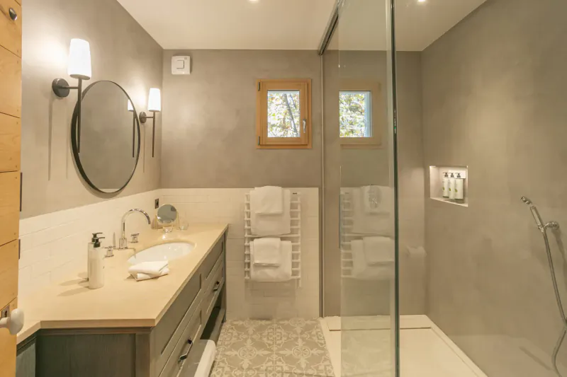 Modern Provence-style bathroom with beige walls, wooden vanity, round mirror, towel warmer, glass shower, and amenities.