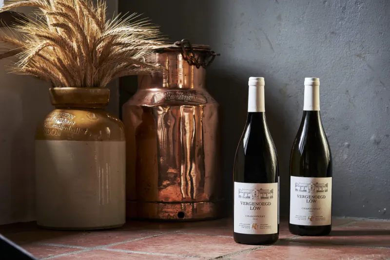 Two bottles of Vergenoegd Löw 'Clara’s Barn' red wine beside copper milk can, terracotta jug, wheat on rustic shelf.
