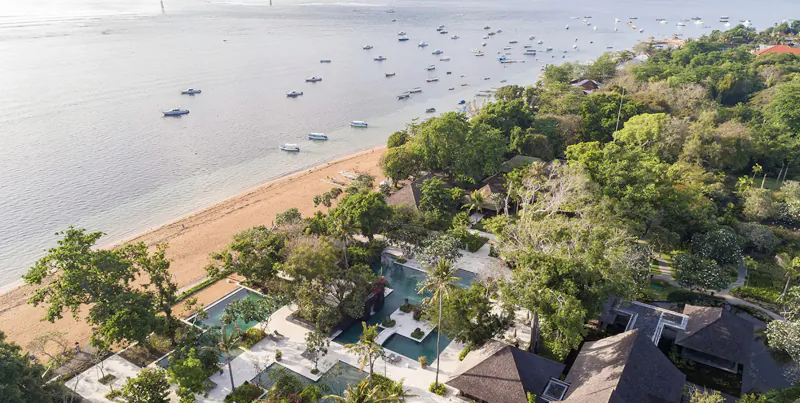 Aerial view of tropical resort with infinity pool, villas, lush gardens, sandy beach, and calm sea with boats.