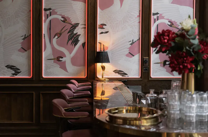 Luxurious hotel gallery bar with pink crane wallpaper, red rose bouquet, brass lamp, velvet chairs, and glassware.