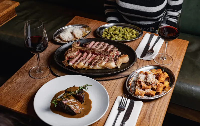 Italian steakhouse table: seared ribeye steak, tagliatelle, peas, rice, red wine glasses on wooden surface.