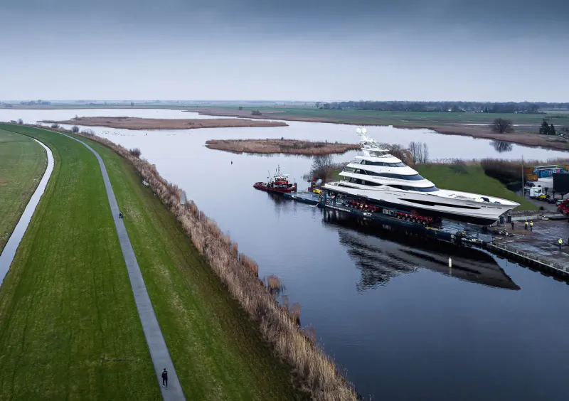 Aerial view of white luxury superyacht docked at canal shipyard with tugboat, surrounded by dikes and wetlands.