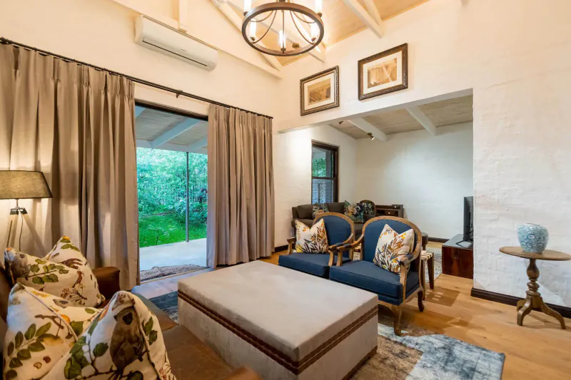 Elegant hotel lounge with blue armchairs, ottoman, lamp, art, and garden view through open doors.