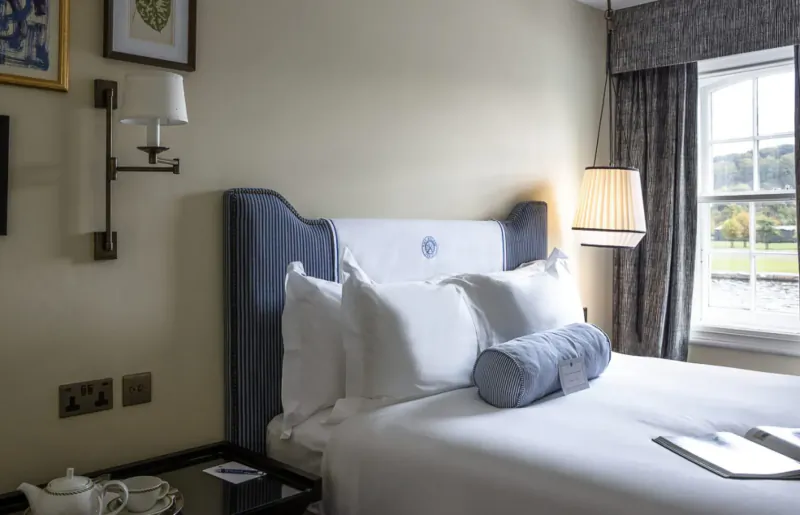 Elegant hotel bedroom with blue upholstered headboard, white pillows, bedside table, and window view of riverside landscape.