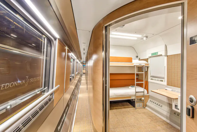 Caledonian Sleeper train corridor with open door to orange bunk beds, large windows, and warm lighting.