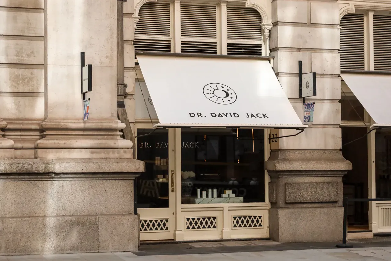 Dr. David Jack storefront with white awning, clock logo, and skincare products in window display on stone building facade.