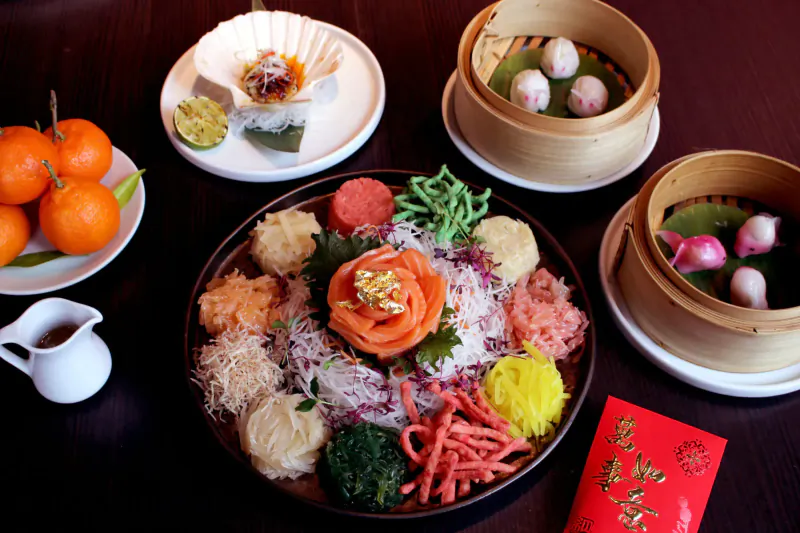 Vibrant Yu Sheng salad with salmon rose, colorful veggies, mandarin oranges, dim sum steamers, and red packet on table