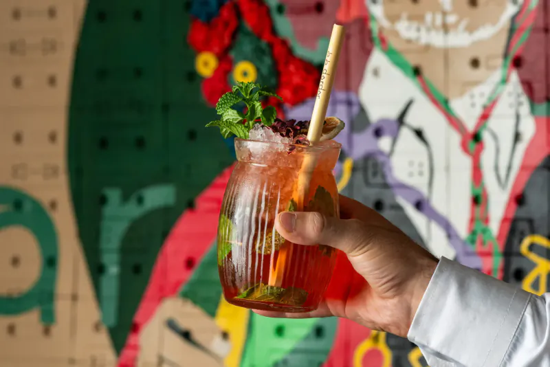 Hand holding tall glass of orange cocktail garnished with mint and berries, bamboo straw, vibrant green graffiti wall background