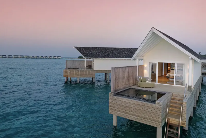 Luxury white overwater villa with open glass doors, lit jacuzzi, and pink sunset sky over turquoise Maldives lagoon
