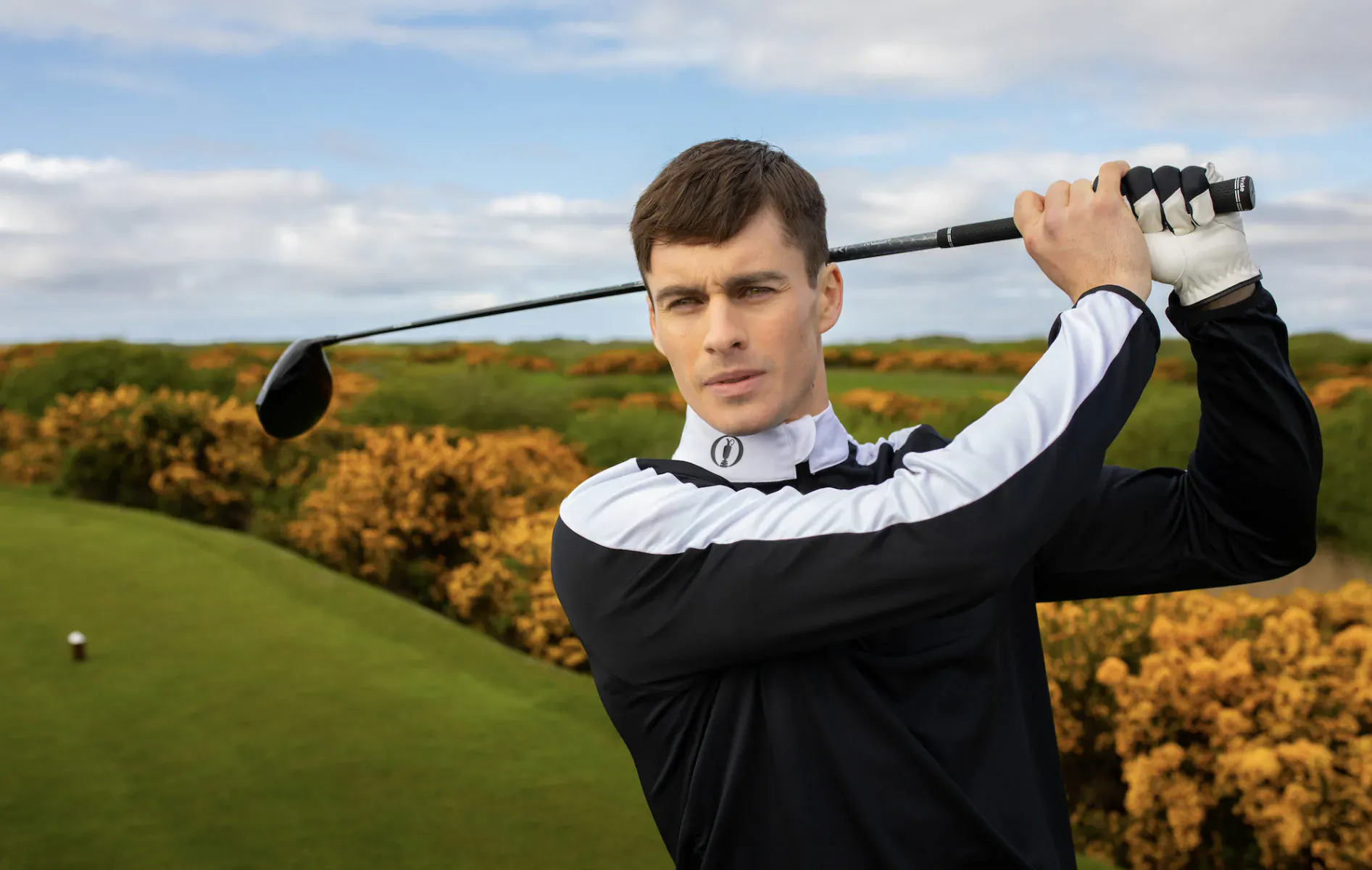 Man in black and white shirt mid-golf swing on green course with yellow gorse and blue sky