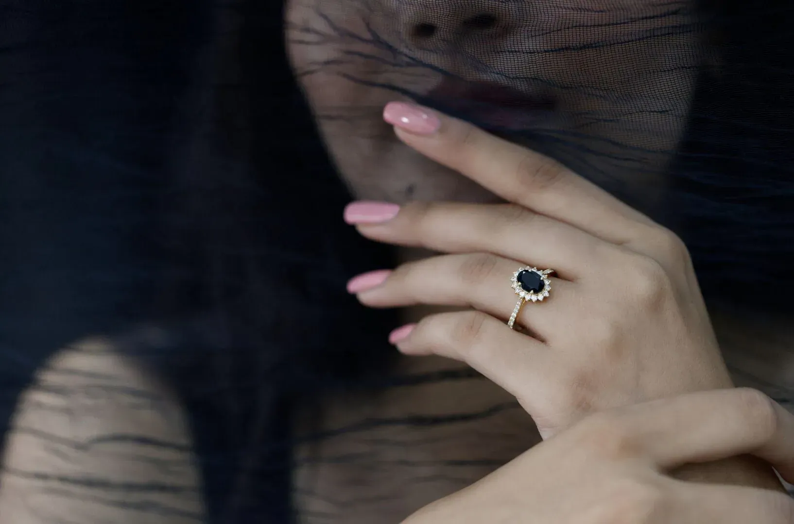 Close-up of woman's hands with pink nails displaying a sparkling black diamond ring, veiled face in background