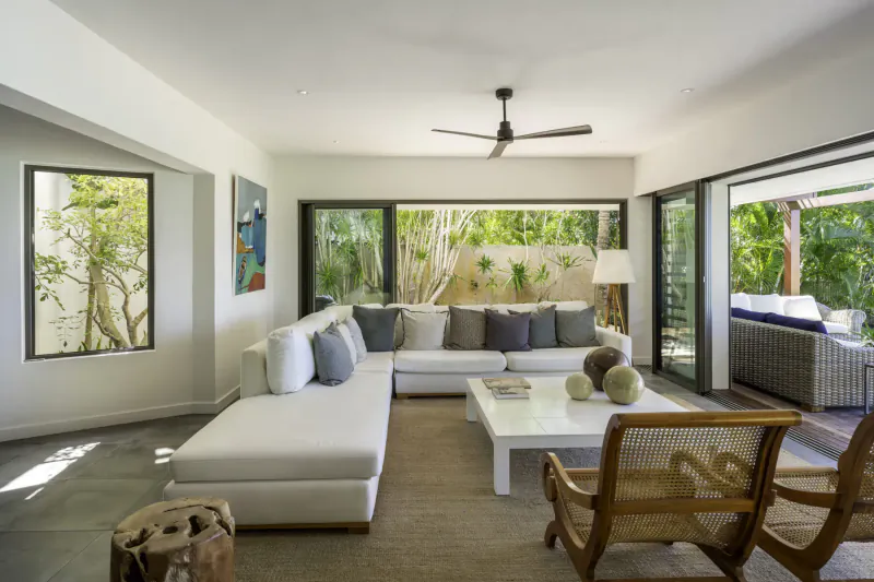 Spacious modern living room in Villa Ruby, Mauritius, with white L-shaped sofa, white coffee table, rattan chairs, ceiling fan, and lush tropical garden views through large sliding doors.
