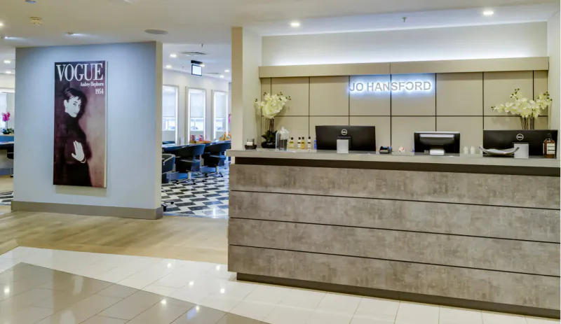 Jo Hansford hair salon reception with Vogue poster, stylish woman image, checkered floor, and neon sign.