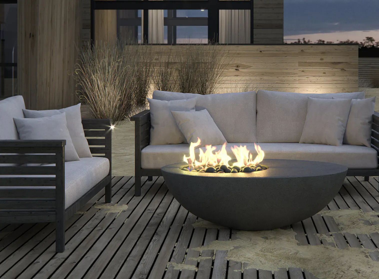 Modern outdoor living area at dusk with beige sofa set around a round black fire pit on wooden deck.