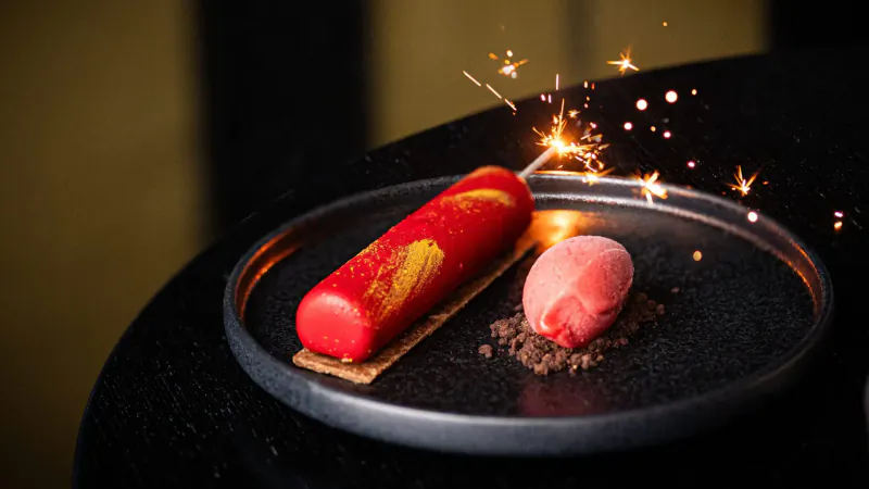 Red entremet with pink ice cream scoop, chocolate crumbs on black plate, lit sparkler, dark setting