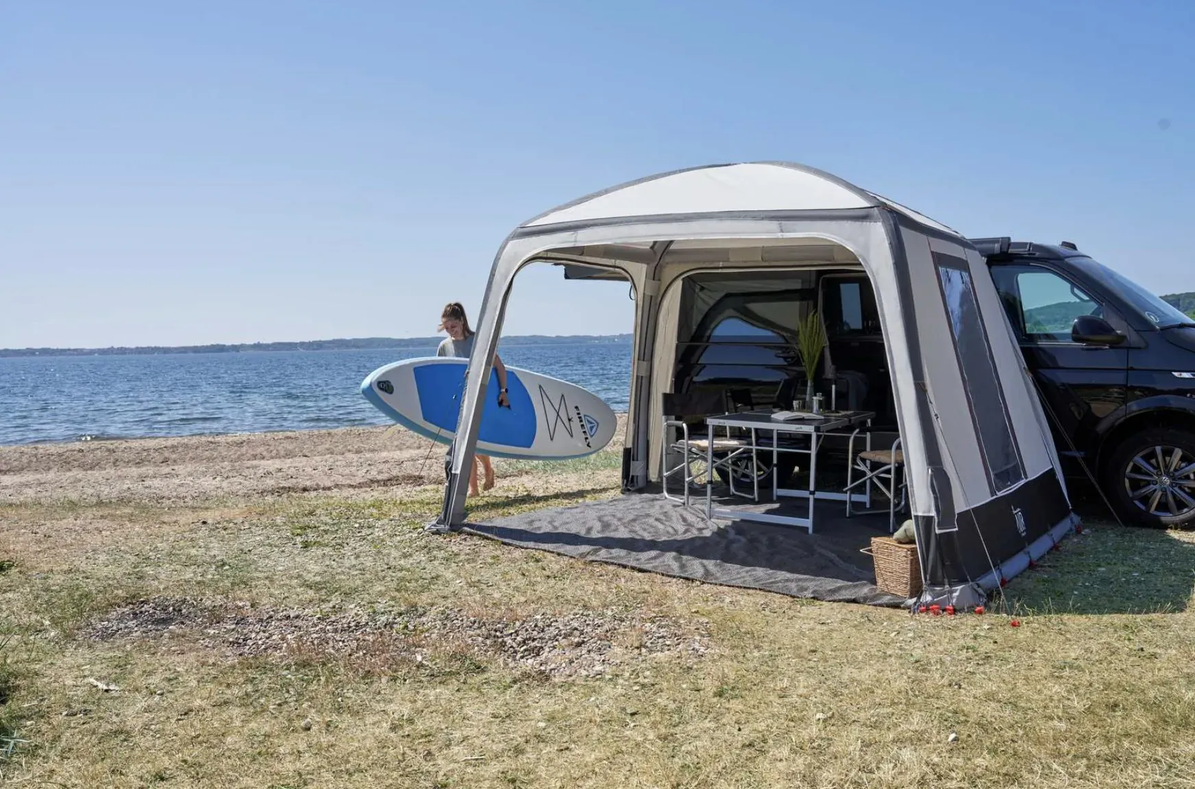 White Isabella awning attached to black motorhome on beach, blue inflatable SUP board and table inside, lake backdrop