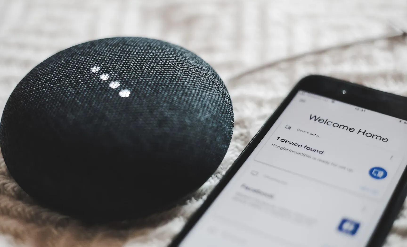Google Home smart speaker beside smartphone displaying 'Welcome Home' on soft fabric surface.