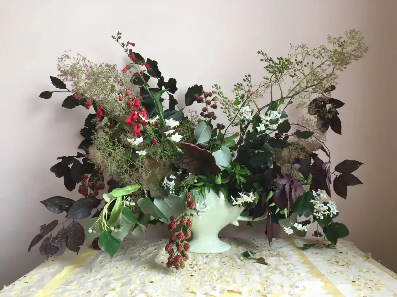 Vibrant floral arrangement in white vase on lace tablecloth, featuring red berries, burgundy leaves, white flowers, and green foliage.
