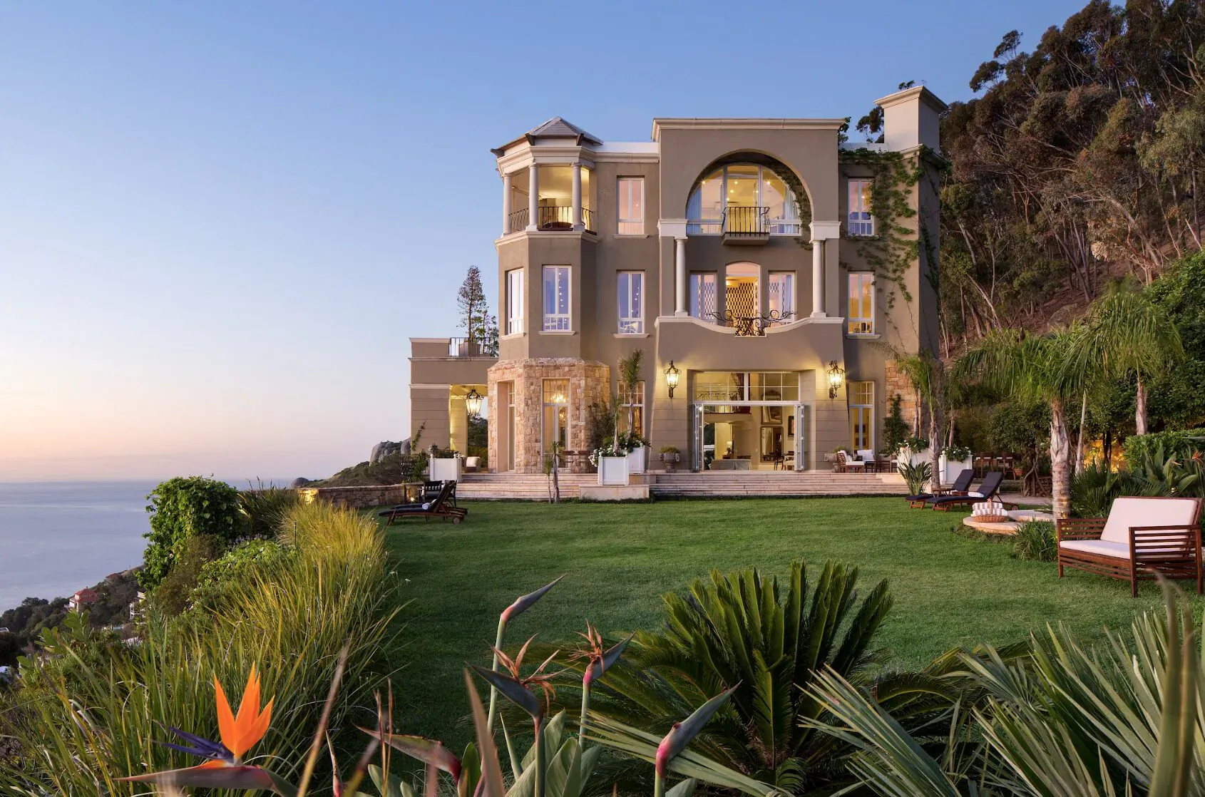 Luxurious beige mansion on grassy cliffside overlooking ocean at sunset, with palms, lounge chairs, and tropical plants.