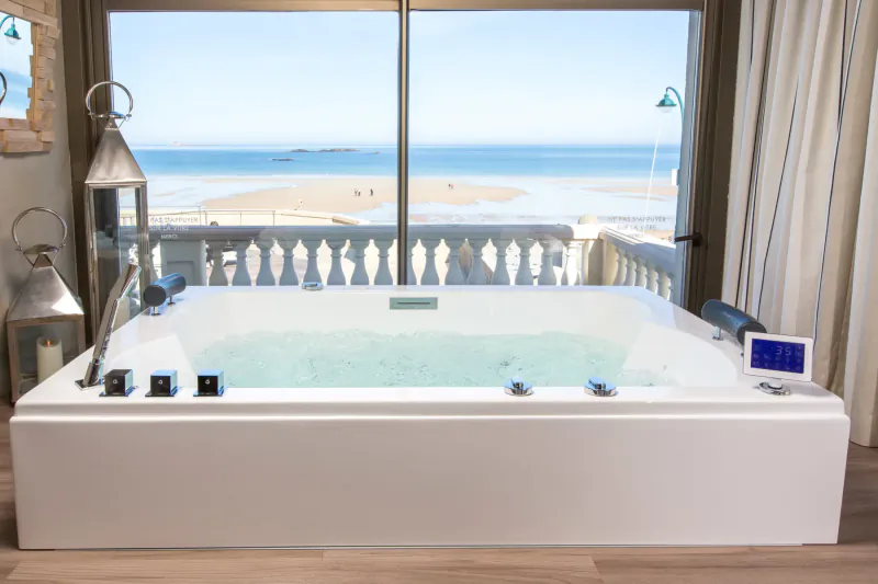 Luxurious white jacuzzi tub in elegant room with sea view to Saint-Malo beach, lanterns and balcony railing.