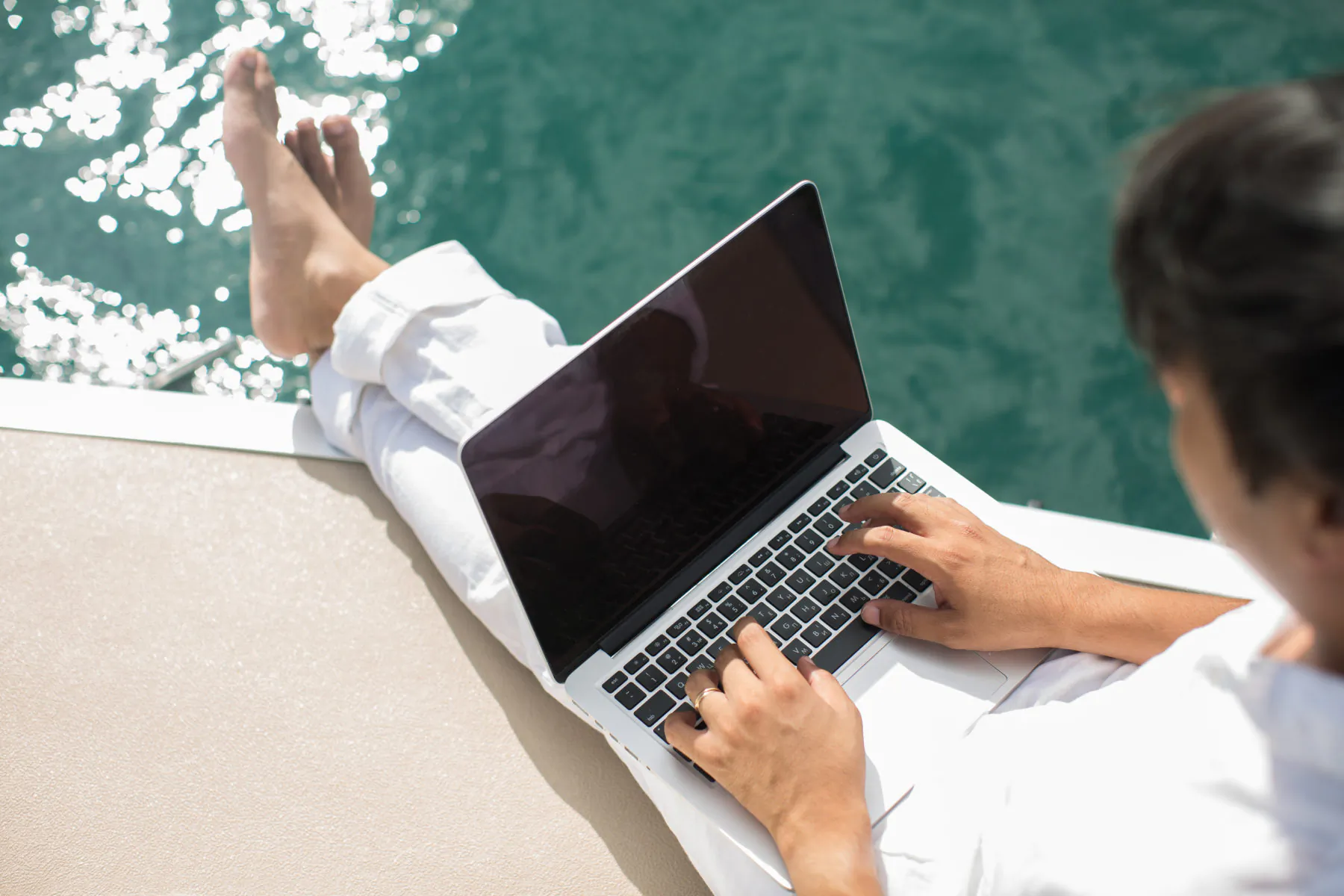 Man in white pants working on laptop, feet dangling over yacht edge into turquoise water