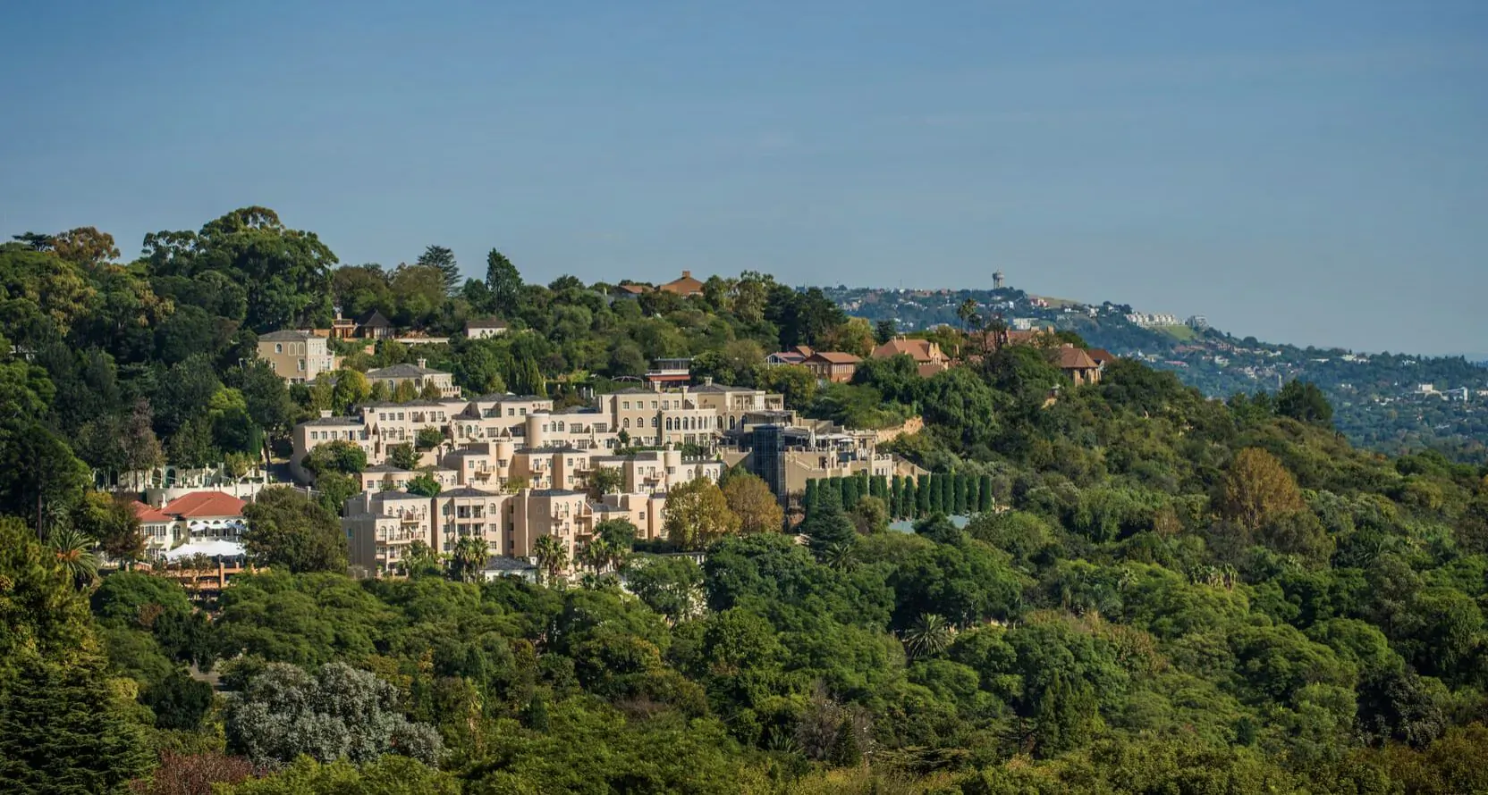 Aerial view of Four Seasons Westcliff hotel amid lush green hills above Johannesburg under blue sky.