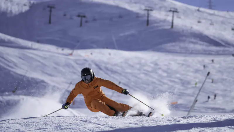 Skier in orange suit and black helmet carving down snowy Pyrenees slope with ski lift in background