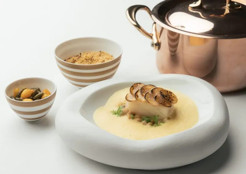 White fish with seared mushrooms on creamy sauce, garnished with herbs, beside striped bowls of chickpeas and grains, copper pot.