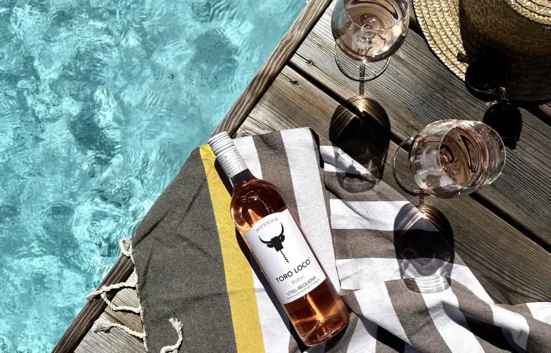 Two glasses of rosé wine 'Torres' bottle on striped towel, straw hat on wooden dock by pool.