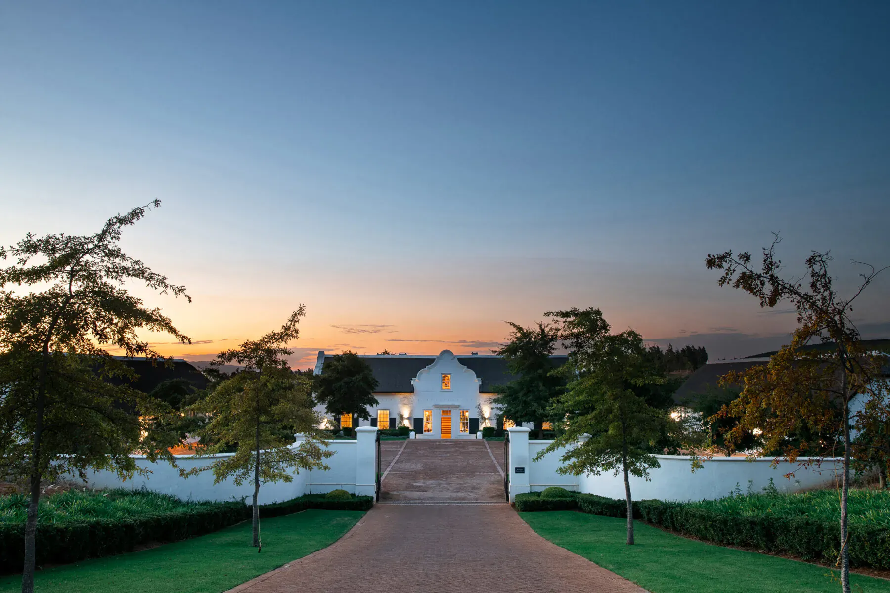 White Cape Dutch manor house at Brookdale Wine Estate at sunset, with palm trees, brick driveway, and white walls.