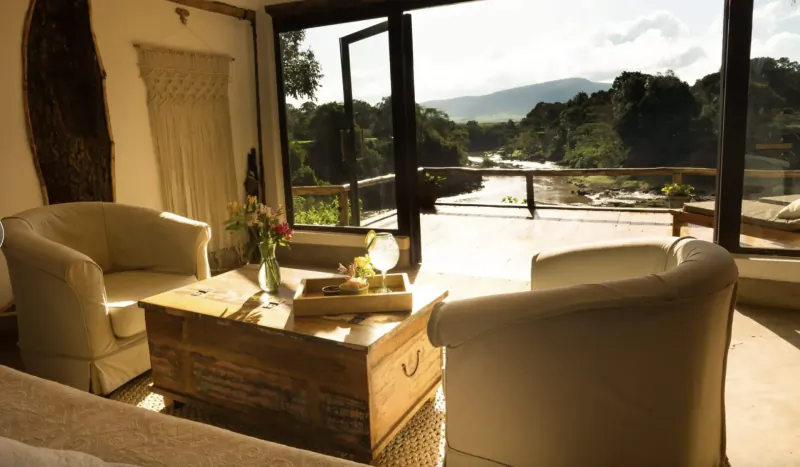 Cozy eco-luxury lounge with cream armchairs, wooden trunk table, flowers, and Maasai Mara river valley view through open doors.