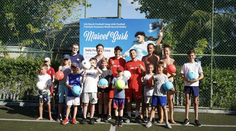 Group of boys and men holding soccer balls, posing in front of 'Marcel Gush' banner on turf field at Lily Beach Resort football camp.