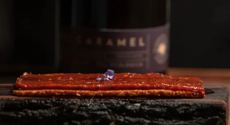 Thin strips of red sauce on charred wooden board with purple flower, Caramelel wine bottle in background