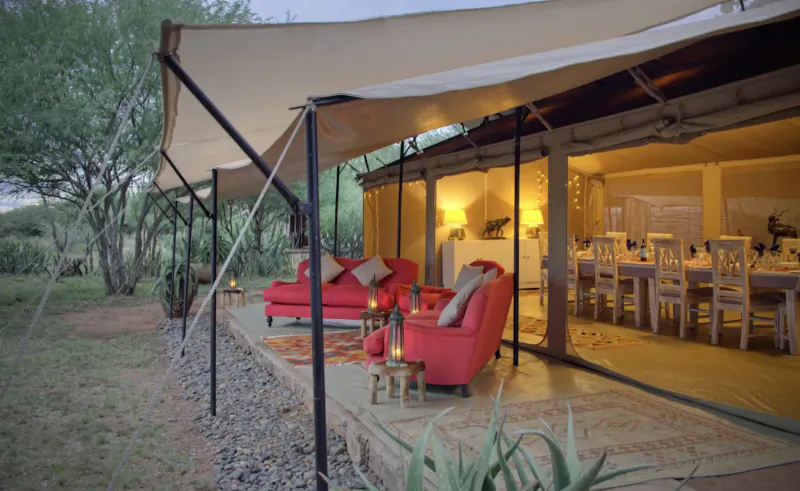 Luxury tent deck with red sofas, lanterns, and dining table inside, African savanna setting at dusk.