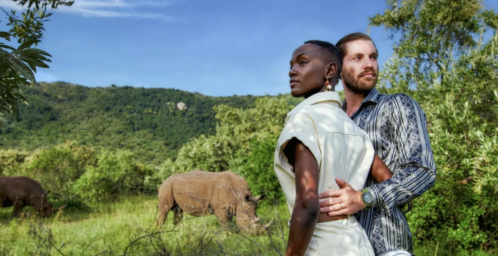 Black woman and bearded man embrace in safari attire, viewing rhinos in lush Mara savanna with hills and trees.