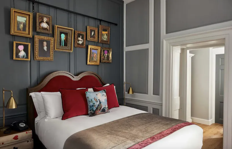 Elegant bedroom with red velvet headboard bed, white pillows, gold quilt, and ornate gilded portrait frames on gray walls.