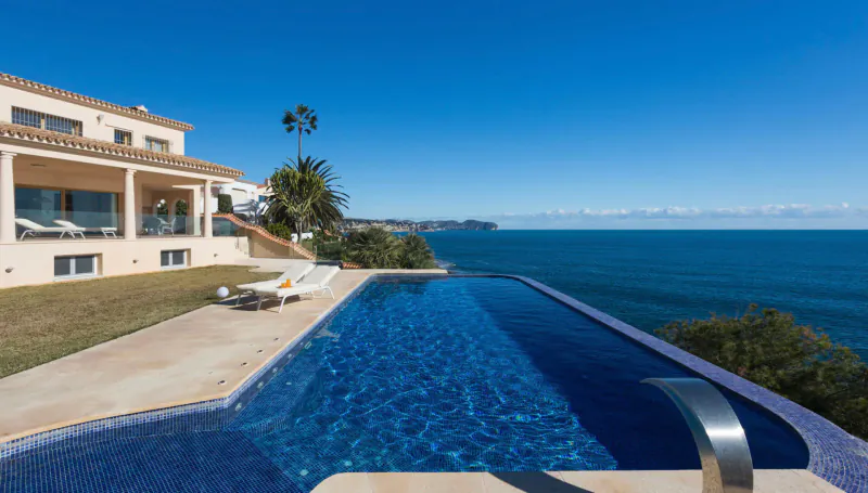 Luxury beige villa with infinity pool overlooking the blue ocean, lounge chairs, palm trees, clear sky.
