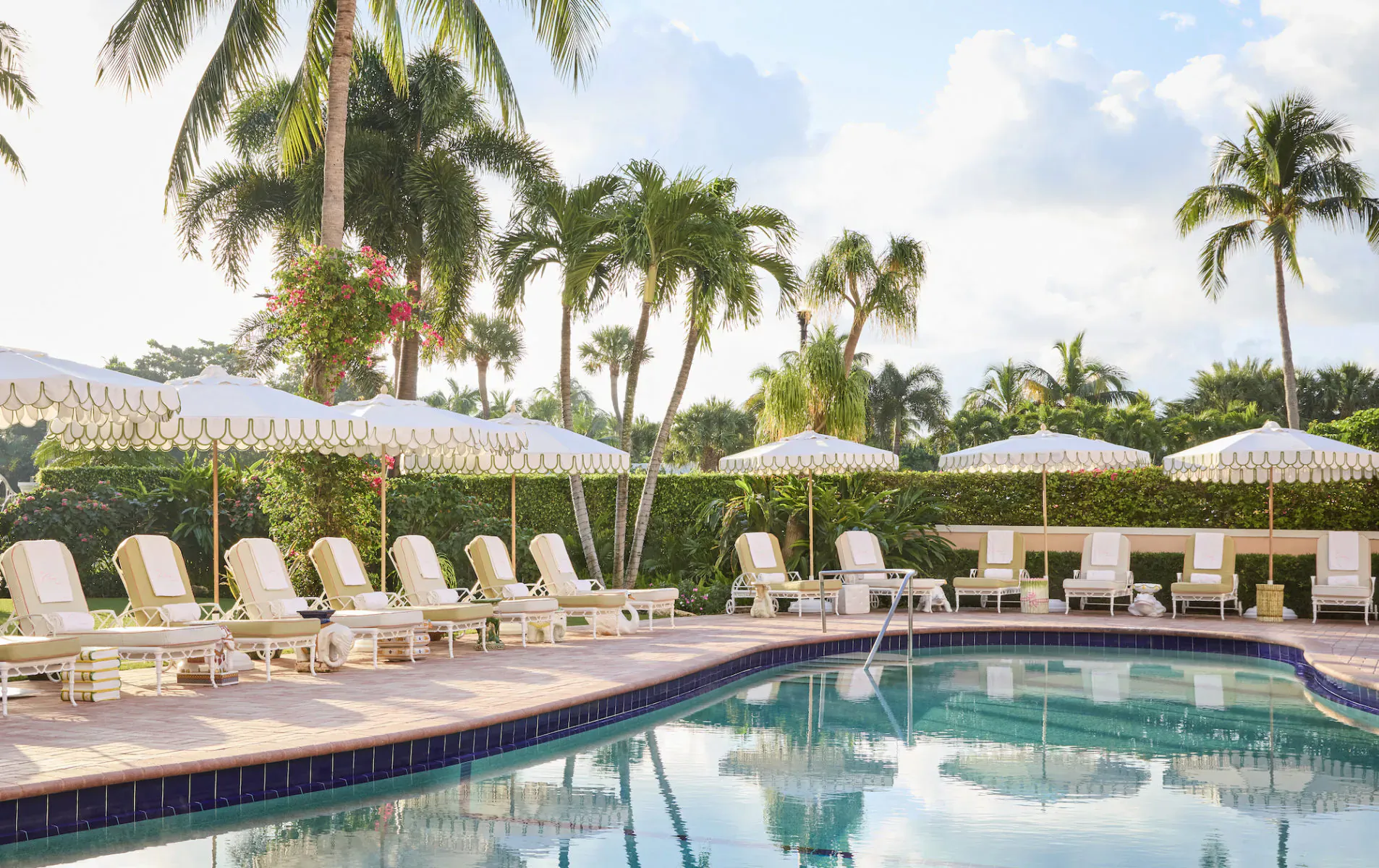 The Colony Hotel's serene Palm Beach poolside with white umbrellas, lounge chairs, palms, and blue water under partly cloudy skies.