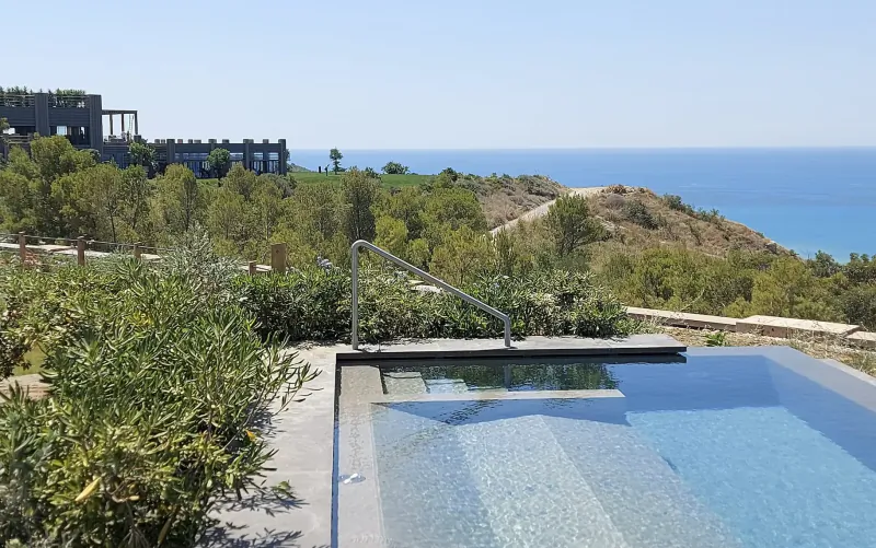 Infinity pool with handrail at ADLER Spa Resort Sicily, overlooking lush greenery, modern lodge, and blue sea.