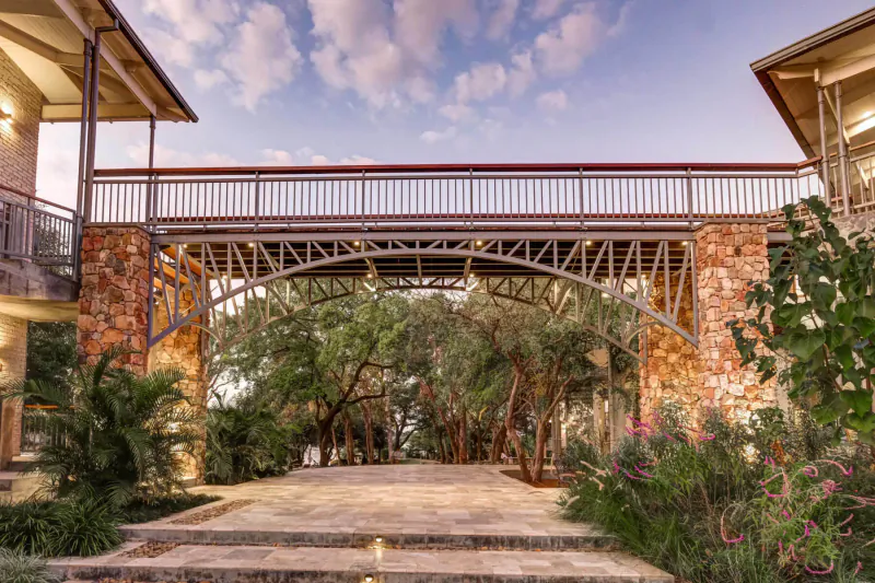 Stone arch bridge spanning lush gardens with trees and palms at Palm River Hotel near Victoria Falls, sunset sky.